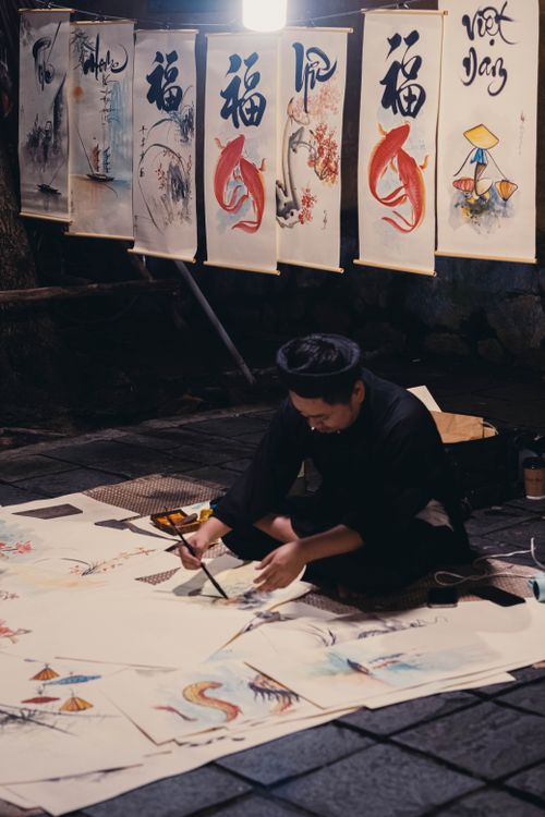 Free An artistic Asian man paints outdoors at night surrounded by traditional art and papers. Stock Photo