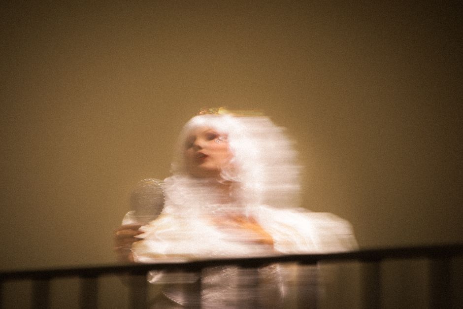 Artistic blurred image of a woman in period costume resembling a ghostly figure on a balcony.