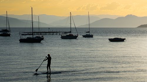 Free A serene scene of a paddleboarder silhouetted against the sunset near a dock with moored sailboats. Stock Photo