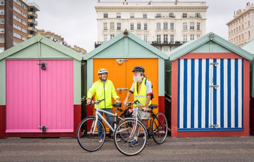 Free Elderly couple enjoying cycling by vibrant beach huts in an urban setting, promoting active lifestyle. Stock Photo