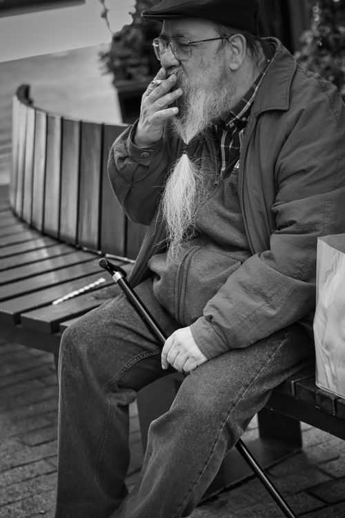Free Black and white portrait of an elderly man smoking while sitting on a bench in the city. Stock Photo