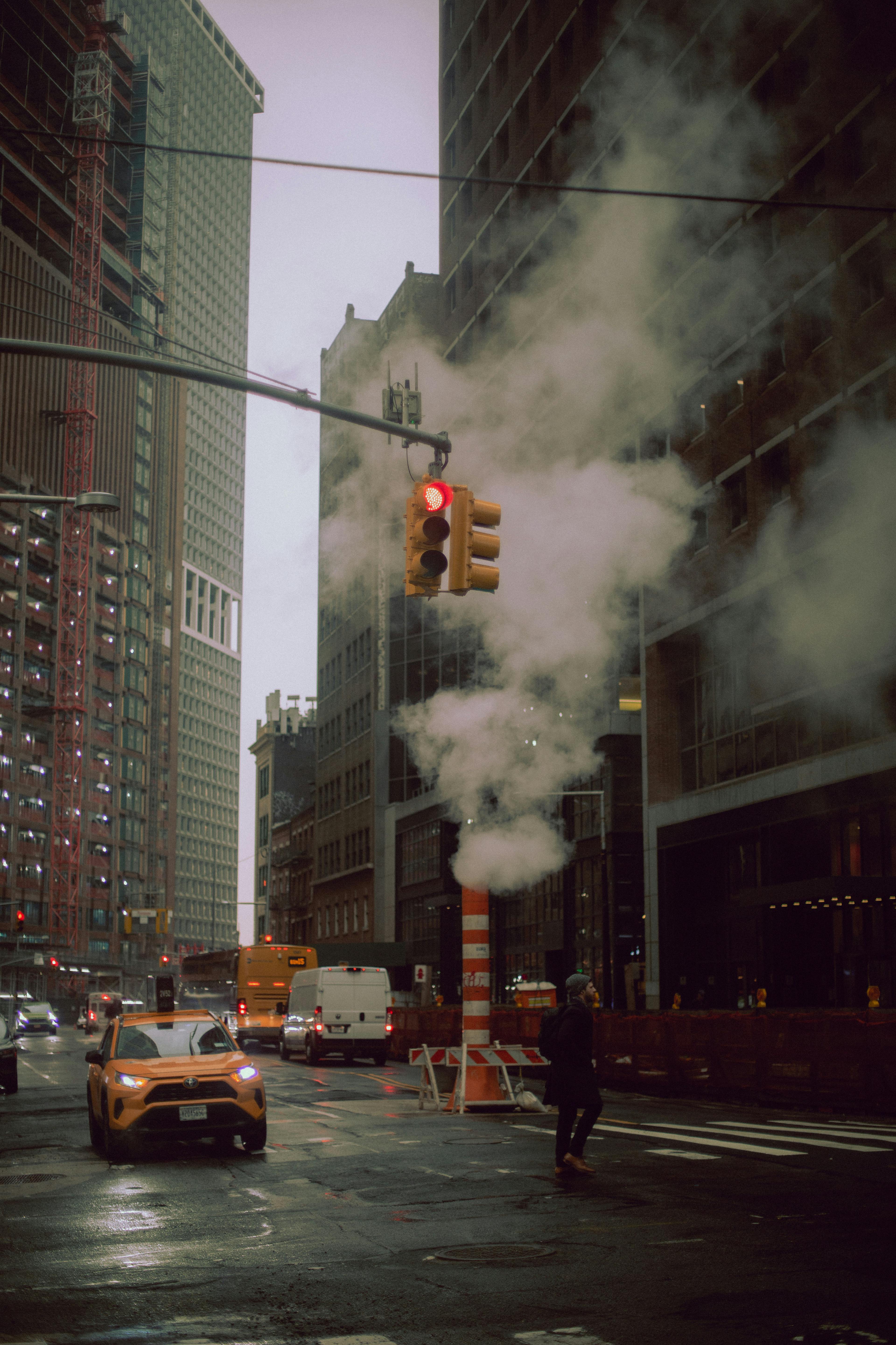 A moody city street scene featuring steam, traffic, and pedestrians under a red light.