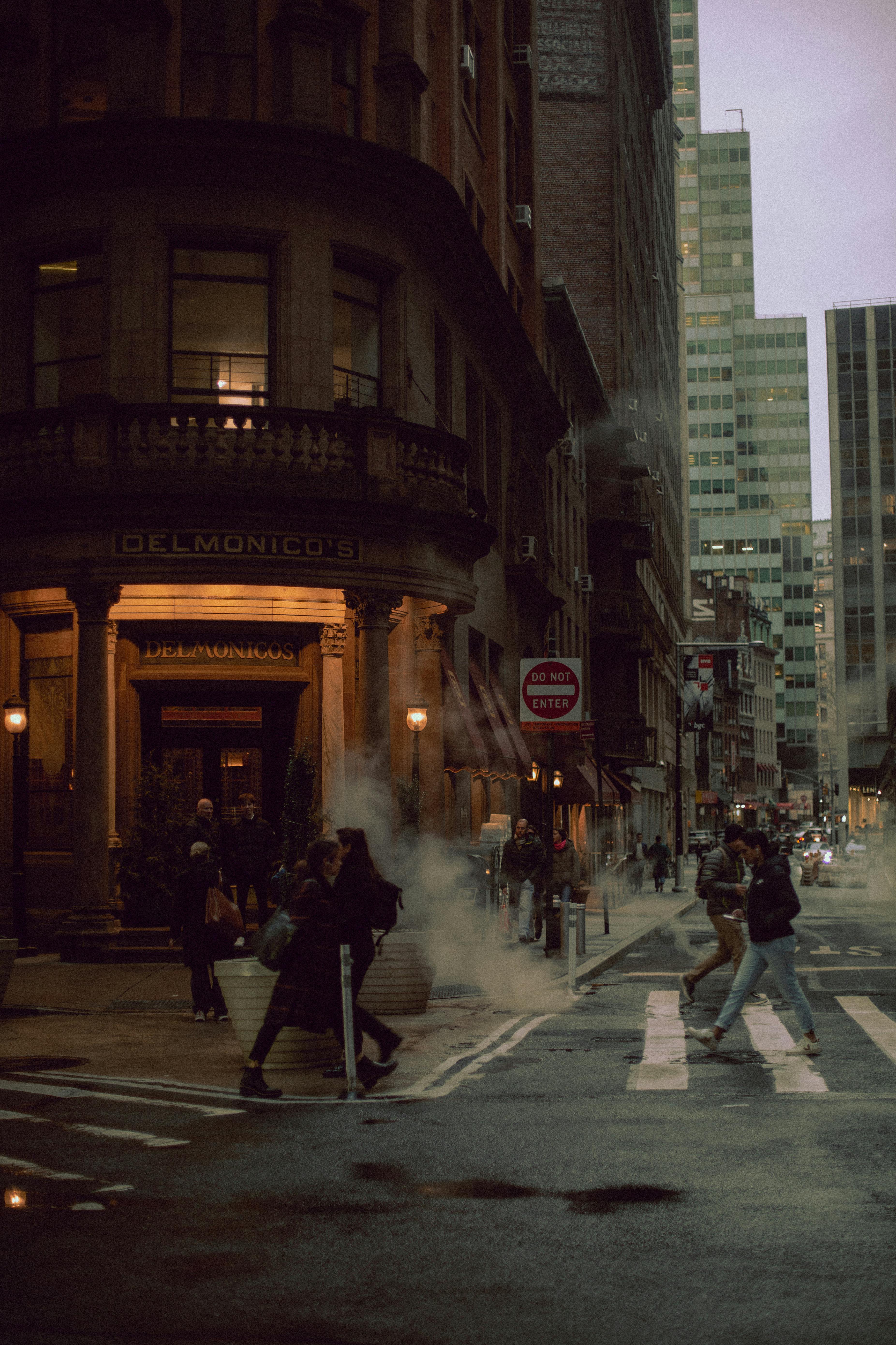 Dusk falls on a bustling city street corner with pedestrians crossing and steam rising.