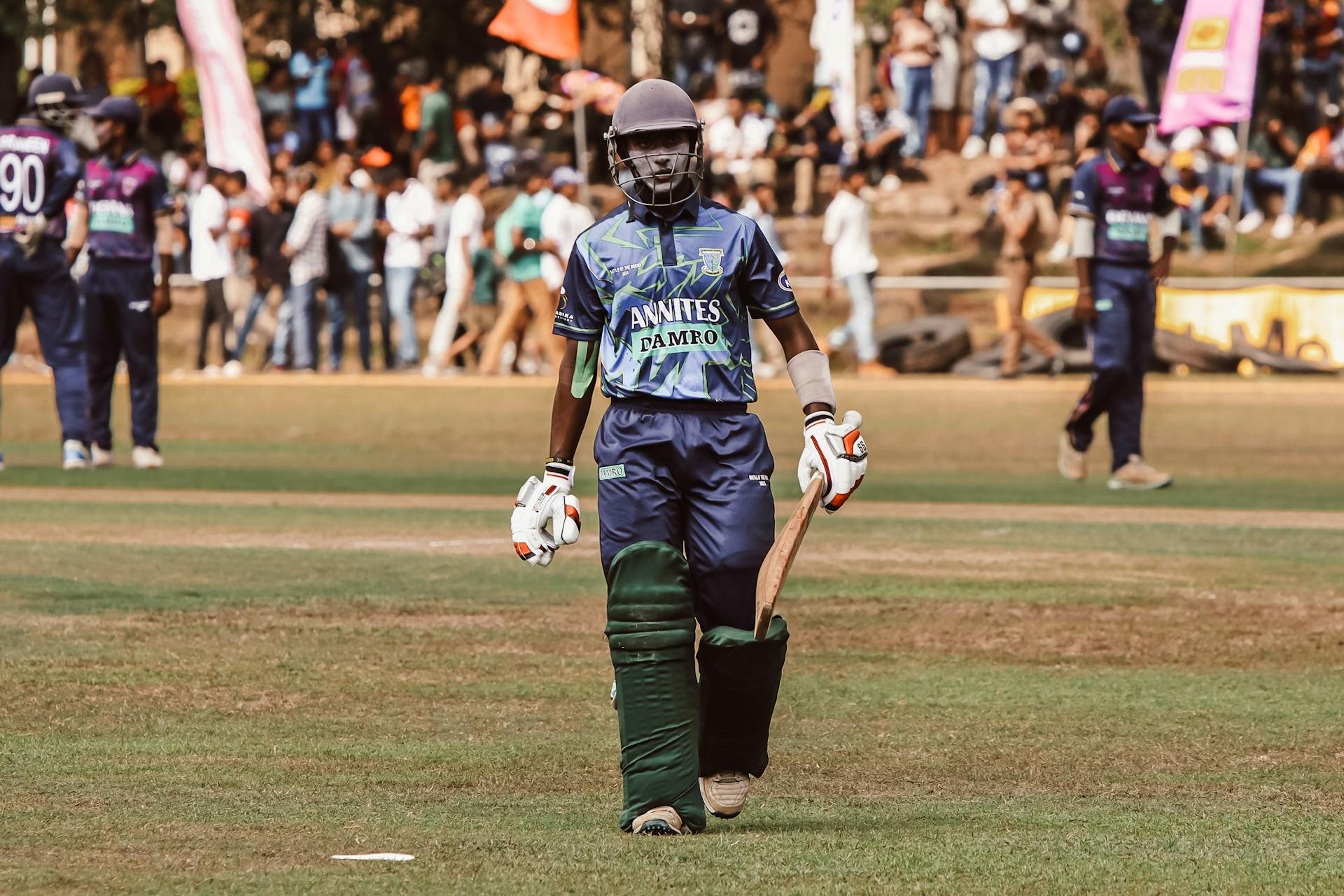 Cricketer wearing protective gear with bat in hand during a match. Crowd and teammates in background.