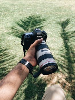 Close-up of a hand holding a DSLR camera with a telephoto lens, outdoors with shadows on grass.