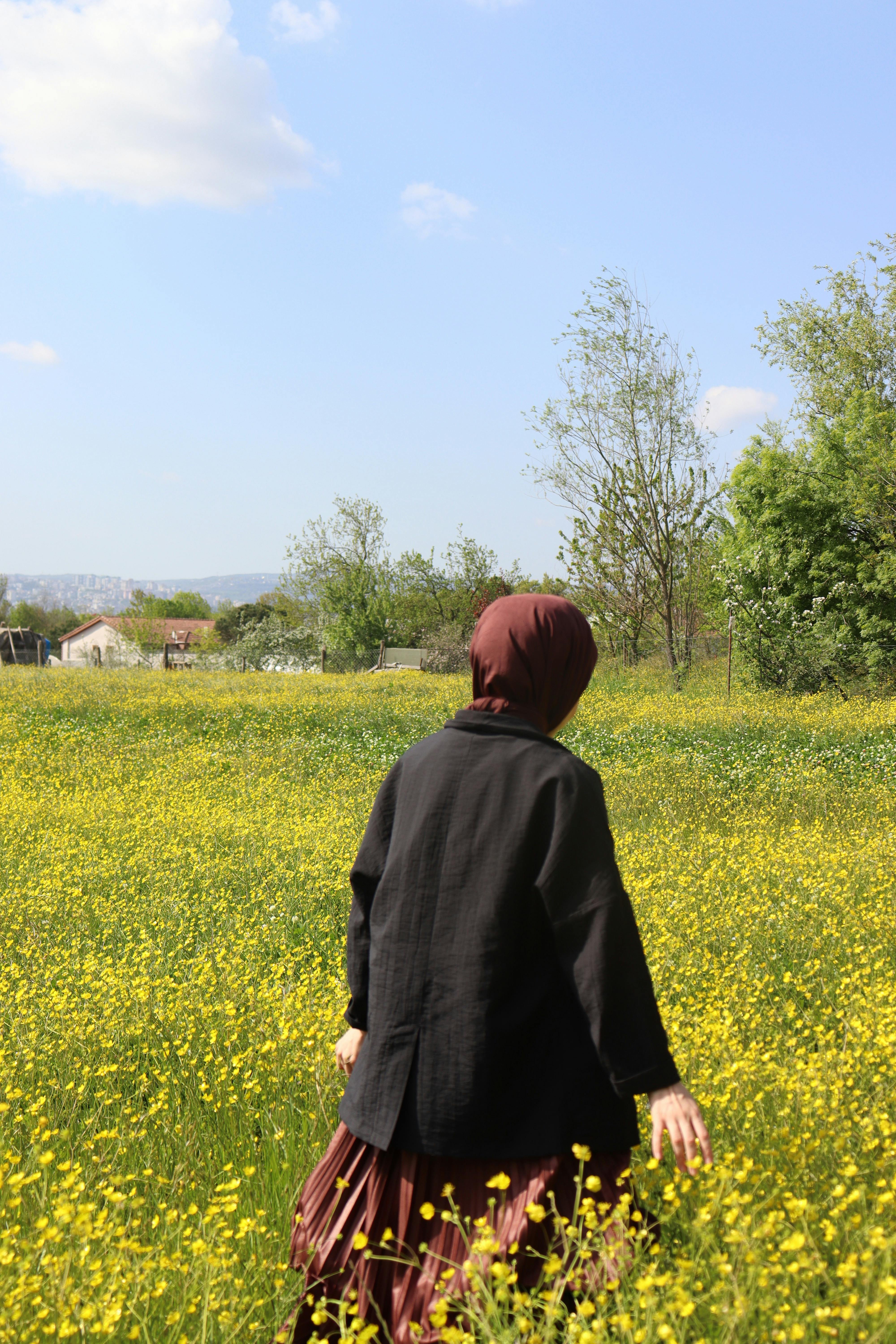A woman in a black jacket and hijab walks through a vibrant meadow of yellow flowers on a sunny day.