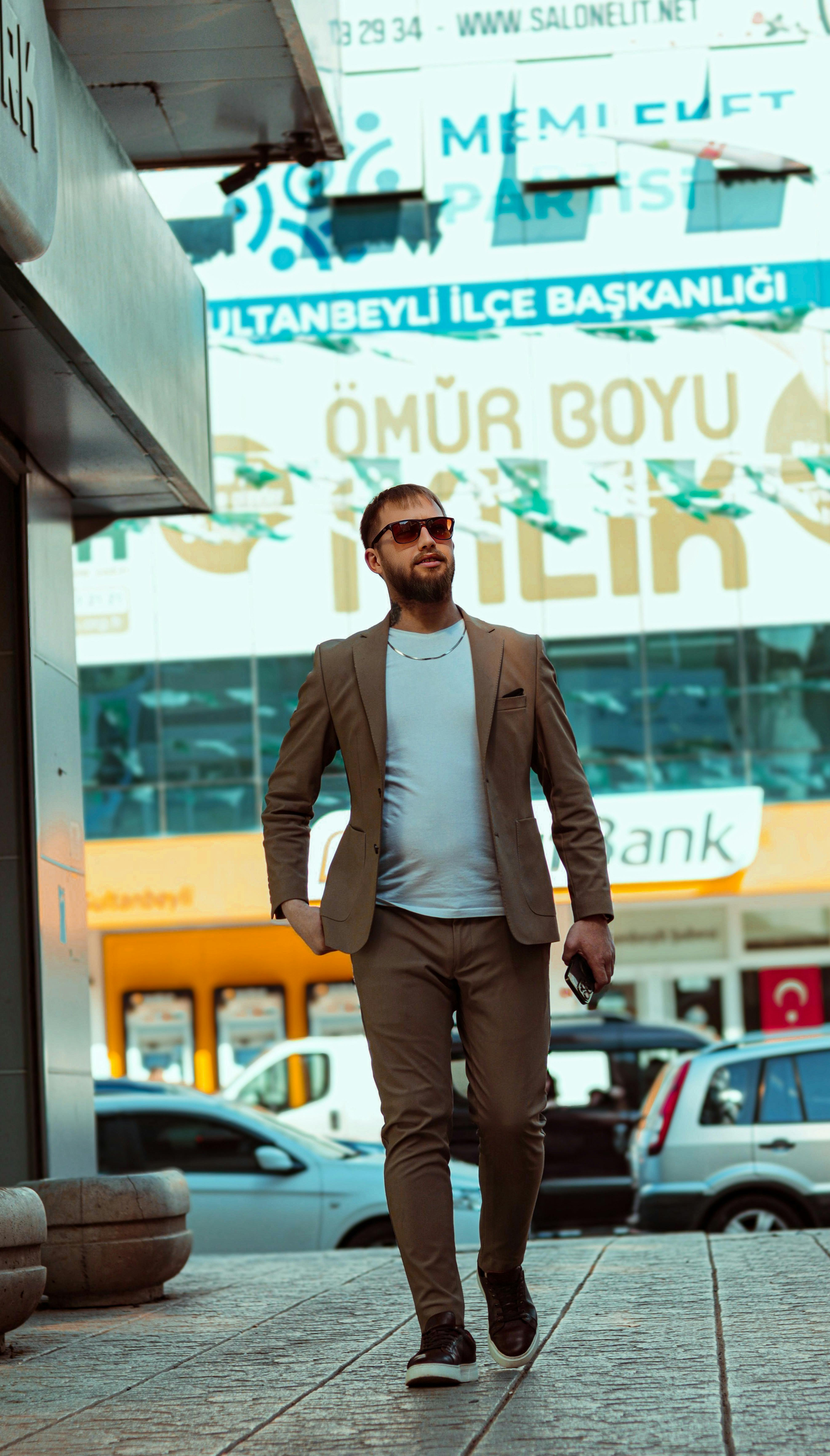 Stylish man in sunglasses and suit jacket walking along urban sidewalk in daylight.