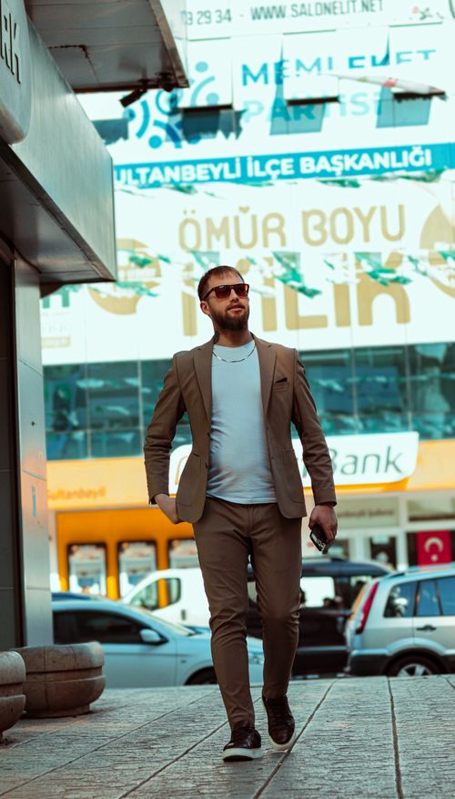 Free Stylish man in sunglasses and suit jacket walking along urban sidewalk in daylight. Stock Photo