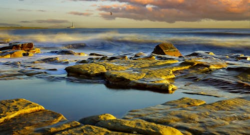 Free View of Waves Crashing on a Rocky Shore and a Distant Lighthouse at Sunset Stock Photo