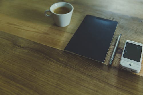 White Ceramic Mug Beside Black Book on Brown Wooden Table