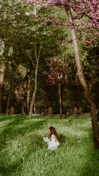 A young girl in a white dress sits among wildflowers in a tranquil park setting.