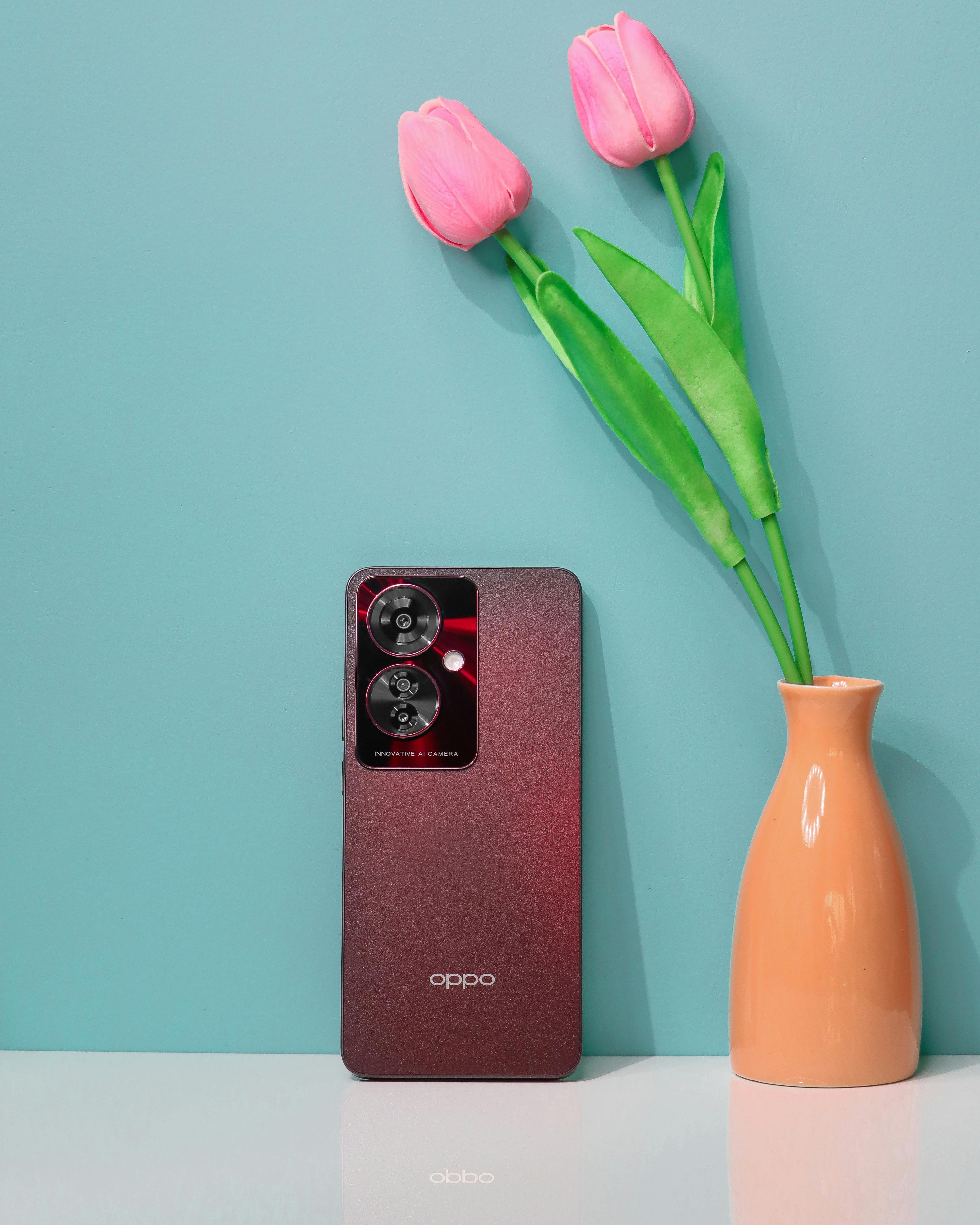 A stylish smartphone next to pink tulips in a vase, set against a blue backdrop.