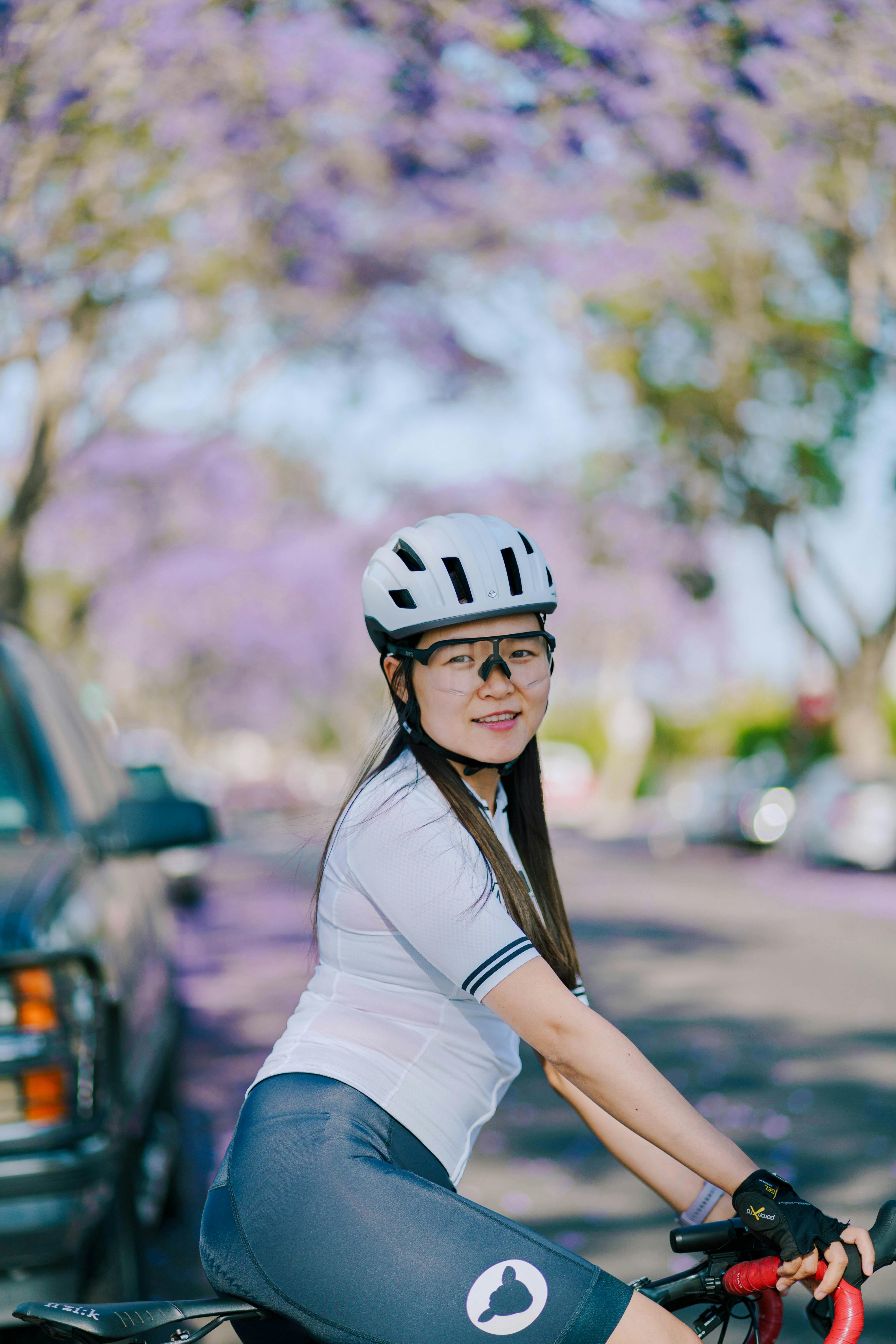 Free A woman in sportswear cycling on a tree-lined street with purple flowers. Outdoor recreation and fitness. Stock Photo