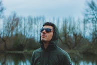 一名男子穿着连帽衫、戴着墨镜，在树木环绕的宁静湖泊附近抬头仰望