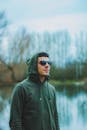 一位时尚男子戴着太阳镜、穿着绿色连帽衫，在宁静的湖边摆姿势，于自然环境中展示风格