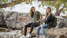 两个十几岁的女孩微笑着坐在水边的木头上，享受户外休闲的一天