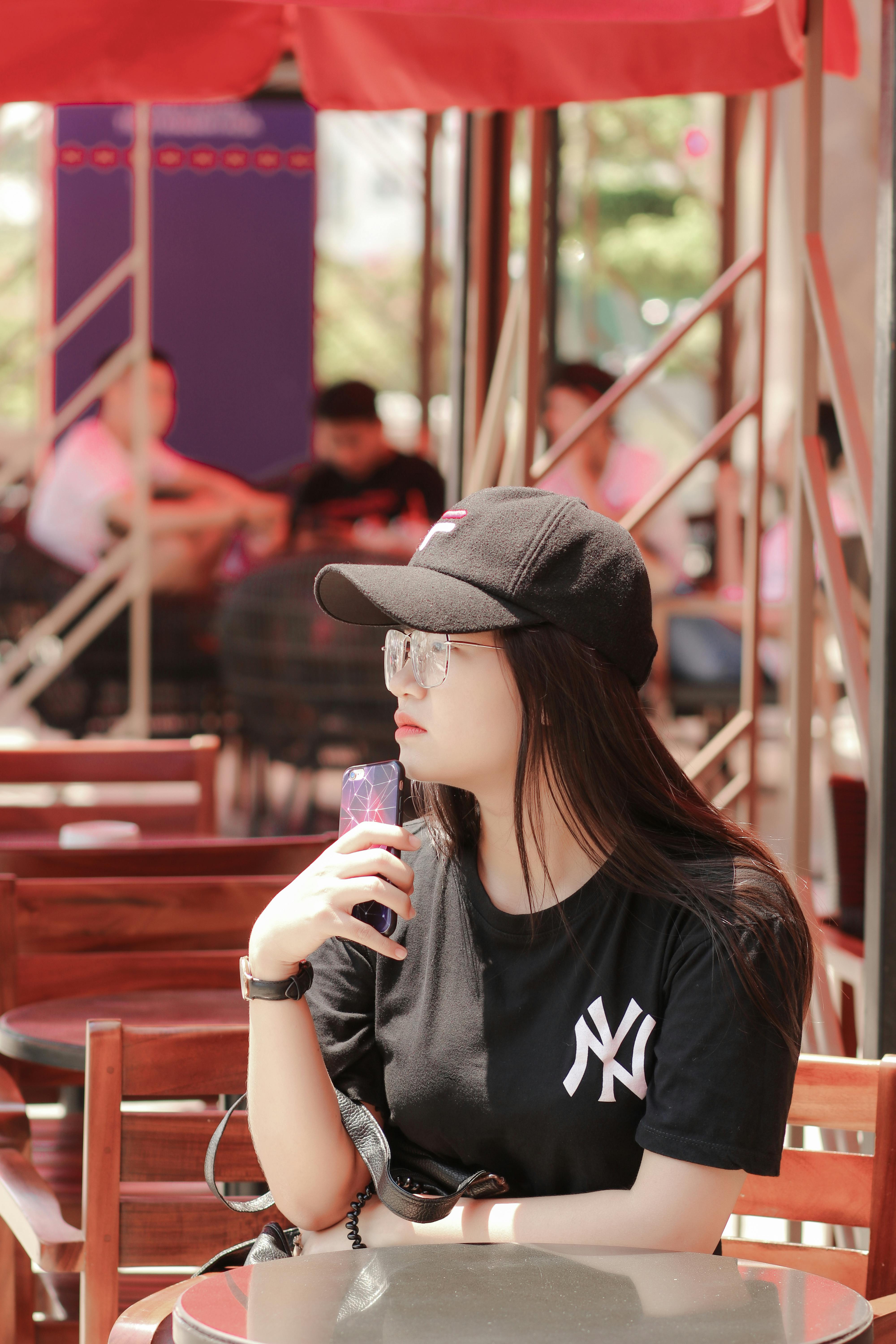 Woman in casual attire with cap holding phone, sitting at an outdoor cafe on a sunny day.