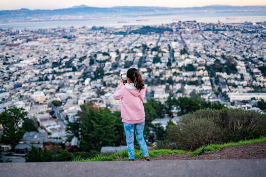 A woman takes a smartphone photo of a sprawling cityscape from a hilltop, showcasing urban beauty.