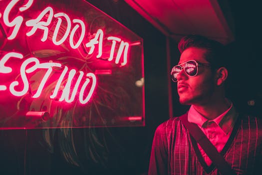 A stylish man wearing sunglasses reflects a vibrant neon sign in an urban night setting.