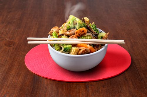 Free A steaming bowl of Asian stir fry with broccoli, carrots, mushrooms, and chopsticks on a red mat. Stock Photo