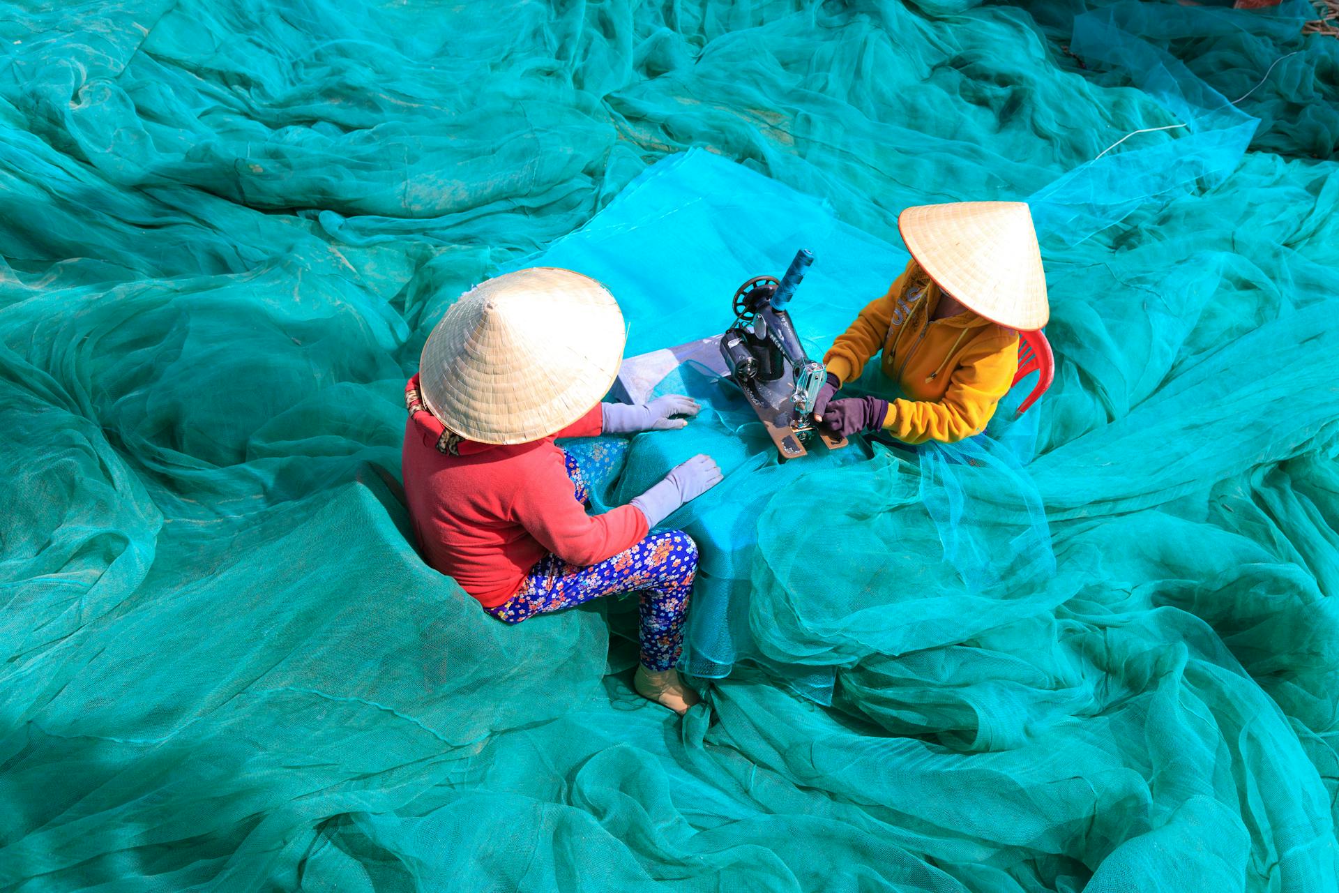 Two women in conical hats repairing fishing nets during daytime. Captured in vibrant colors.