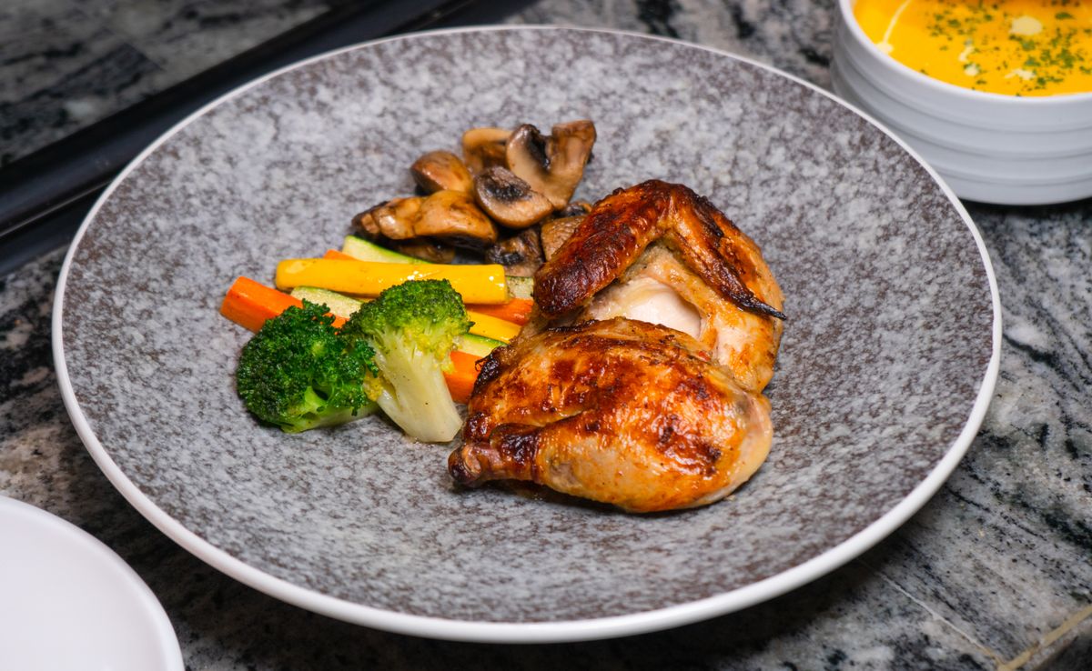 Roasted half chicken with glossy skin beside sauteed mushrooms, broccoli, carrots and squash on gray plate