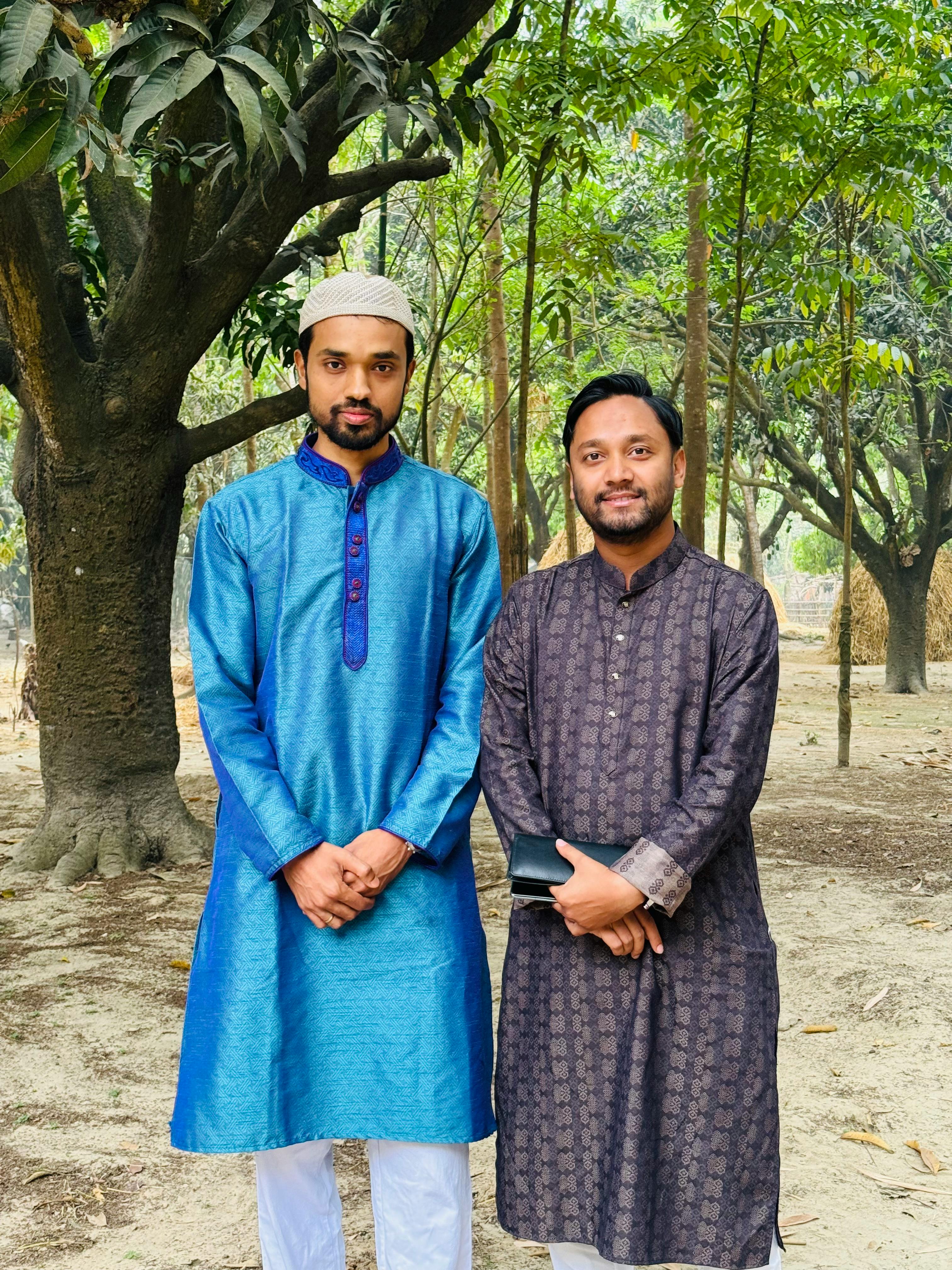 Two men in traditional clothing stand in a forest setting, showcasing cultural attire.