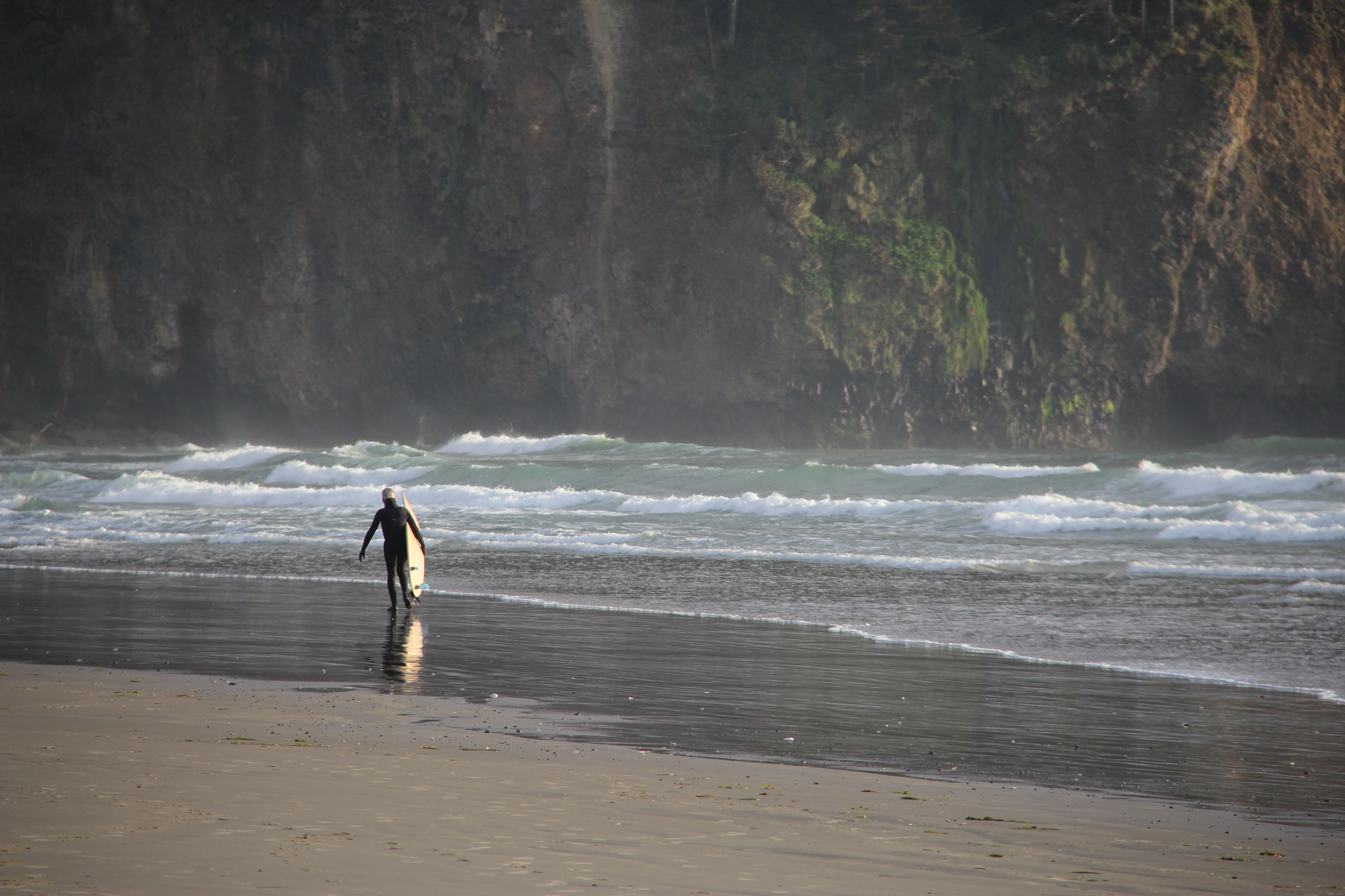 A surfer carrying a board walks along Cape Meares beach with waves crashing and cliffs in the background.