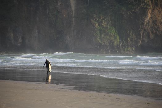 A surfer carrying a board walks along Cape Meares beach with waves crashing and cliffs in the background.