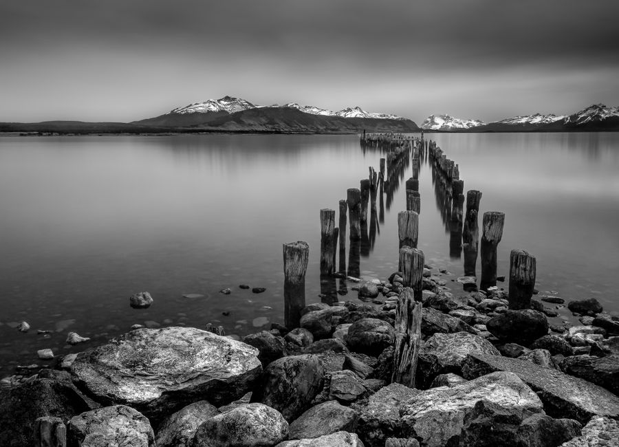 Tranquil black and white photograph of wooden posts along the Puerto Natales coastline with distant mountains.