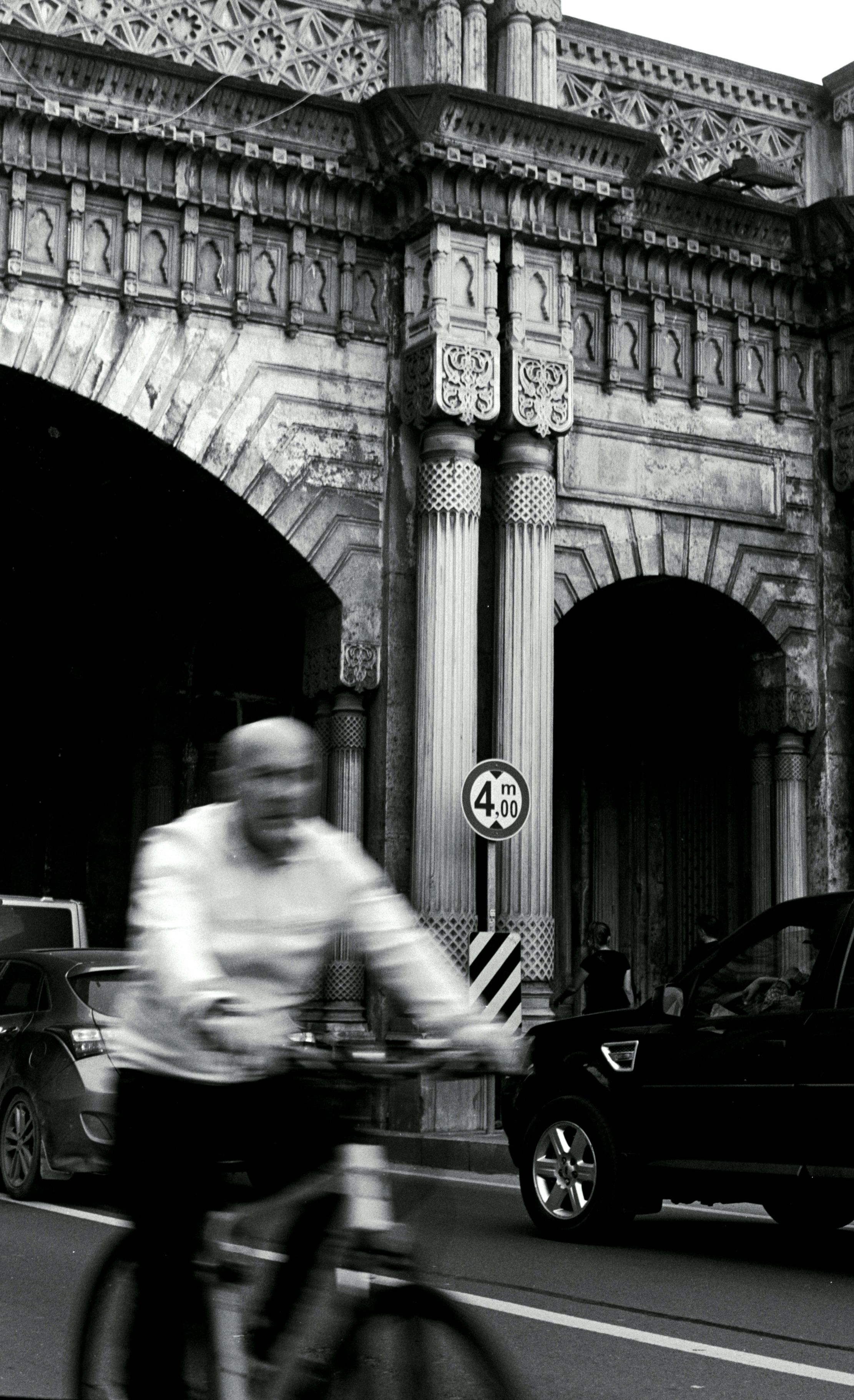 Black and white image of a cyclist in motion near an ancient stone archway in the city.