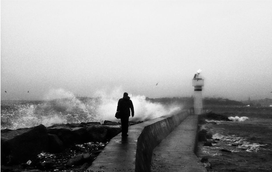 A lone man walks towards a lighthouse on a stormy pier in black and white.
