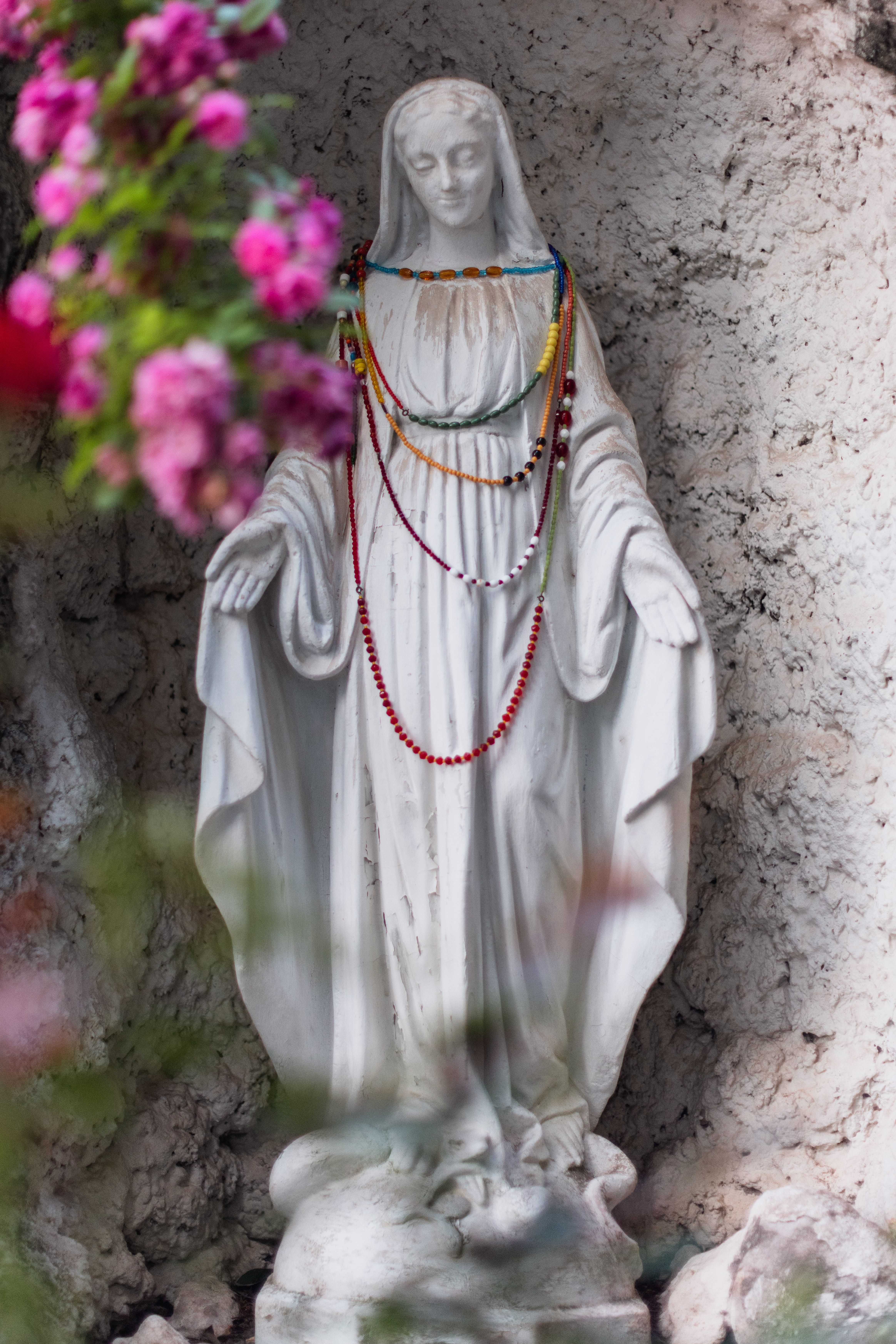 White stone Virgin Mary statue decorated with colorful beads surrounded by pink flowers.