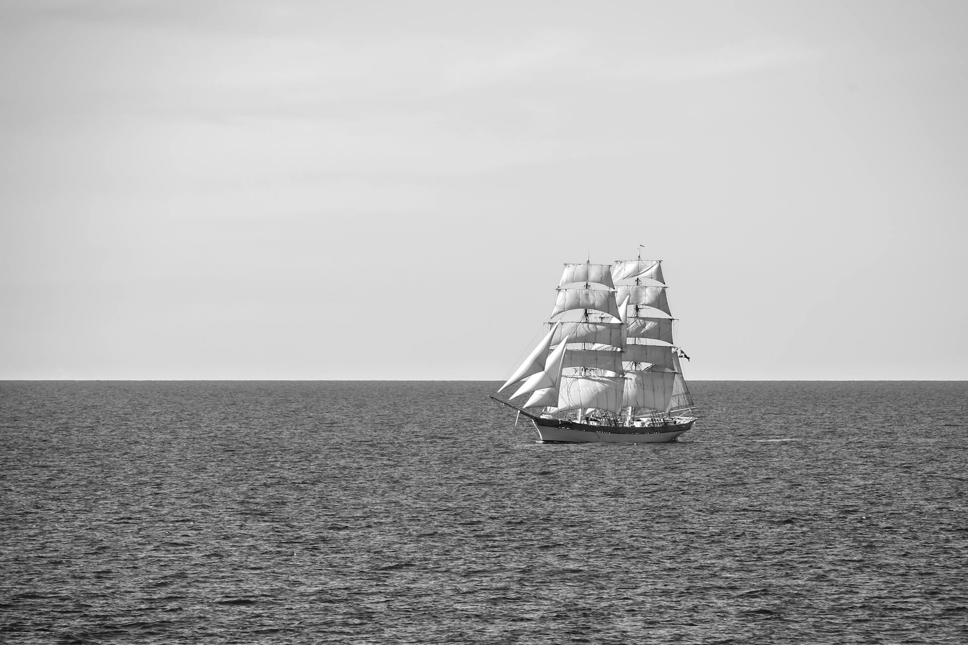 Black and white photo of a sailing ship on a calm ocean, showcasing nautical adventure.