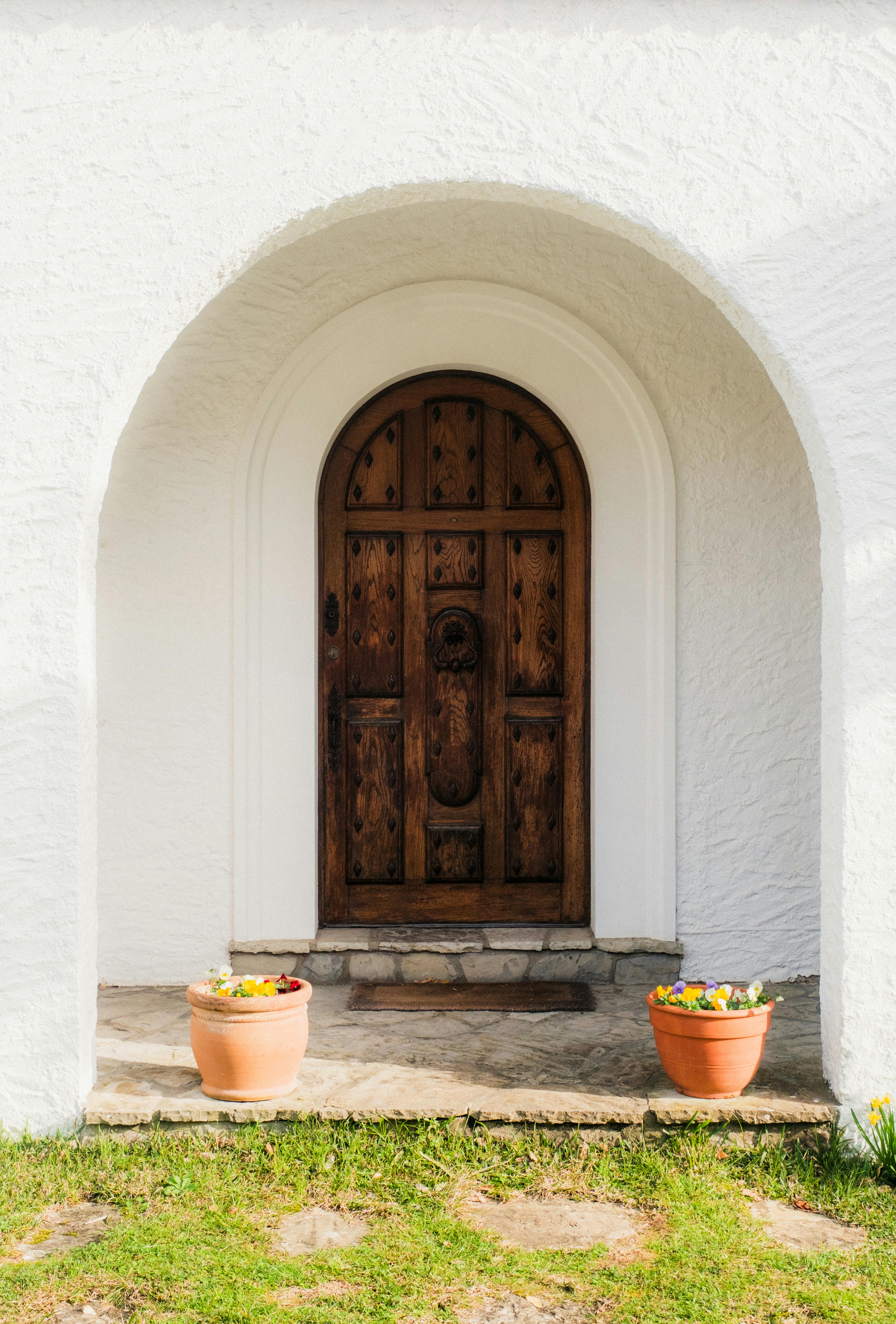 A beautiful rustic wooden door set within a white archway, flanked by potted plants and a garden path.
