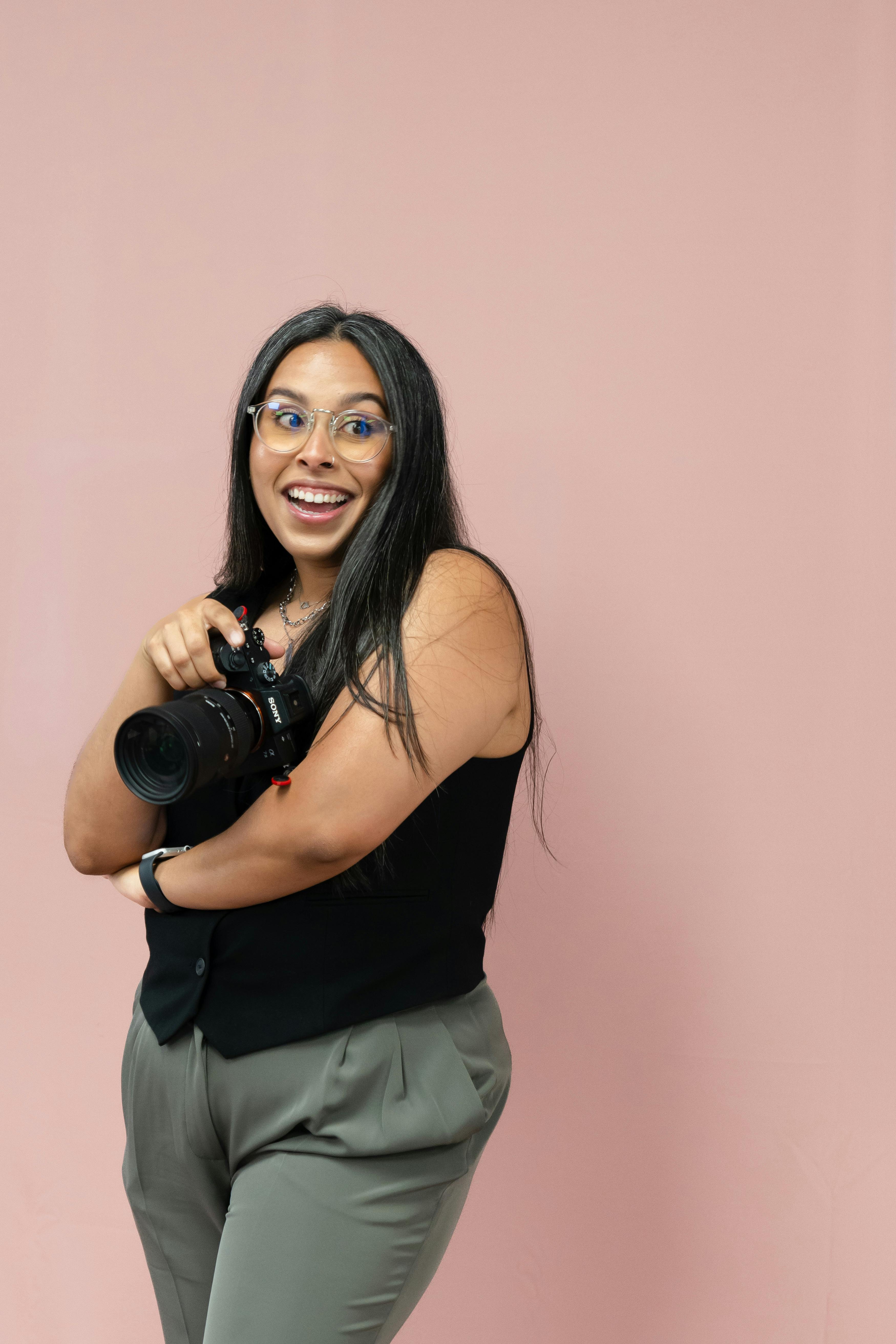 Smiling woman holding a camera in a studio with a pink background.