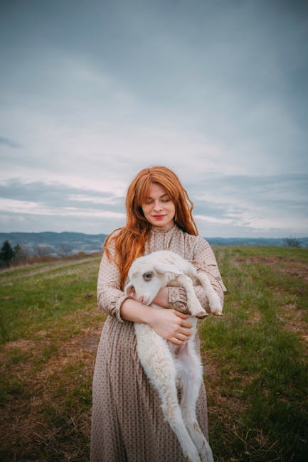 A red-haired woman in a dress holds a lamb in a tranquil outdoor setting under a cloudy sky.