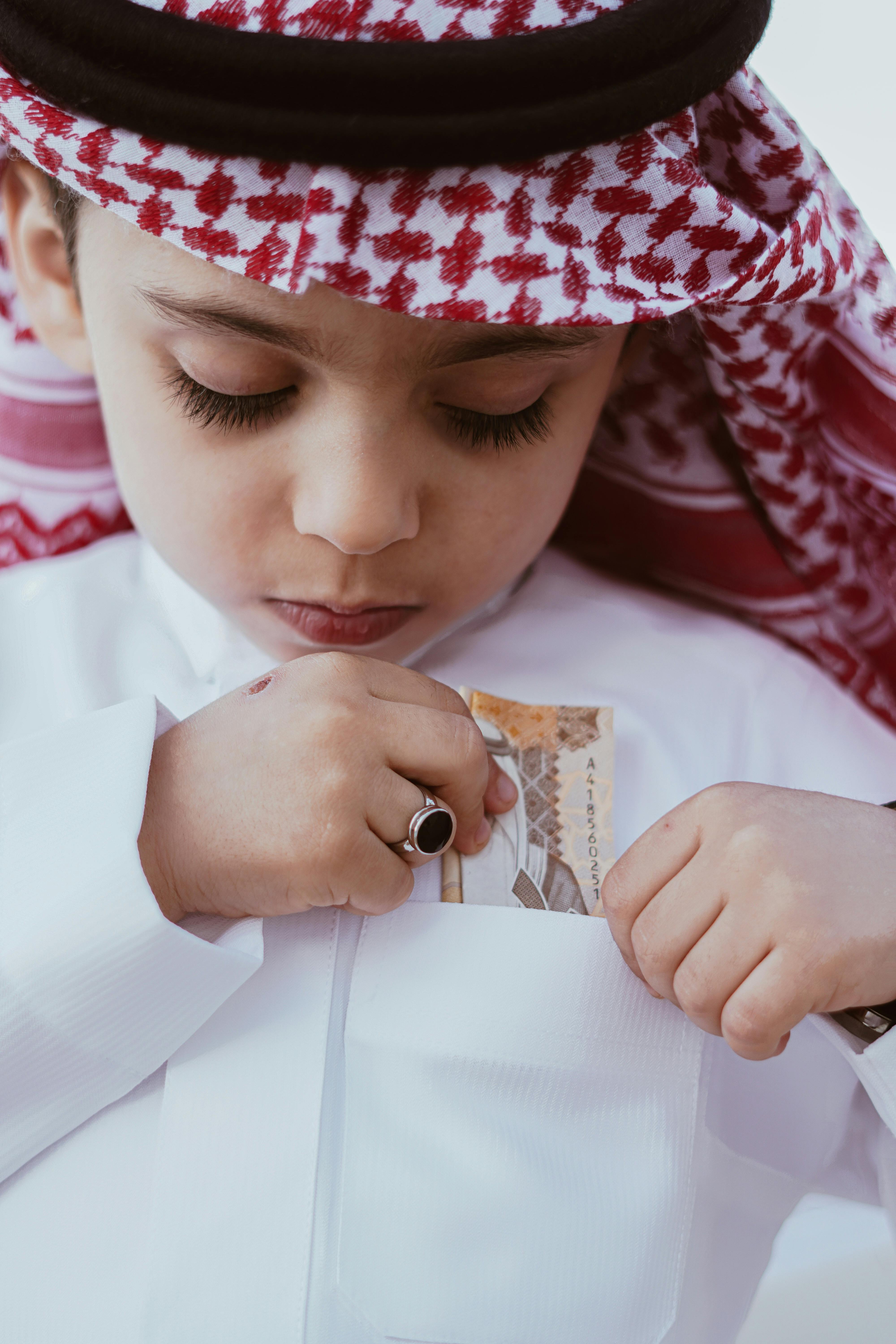 A young boy in traditional Middle Eastern attire putting a banknote in his pocket.