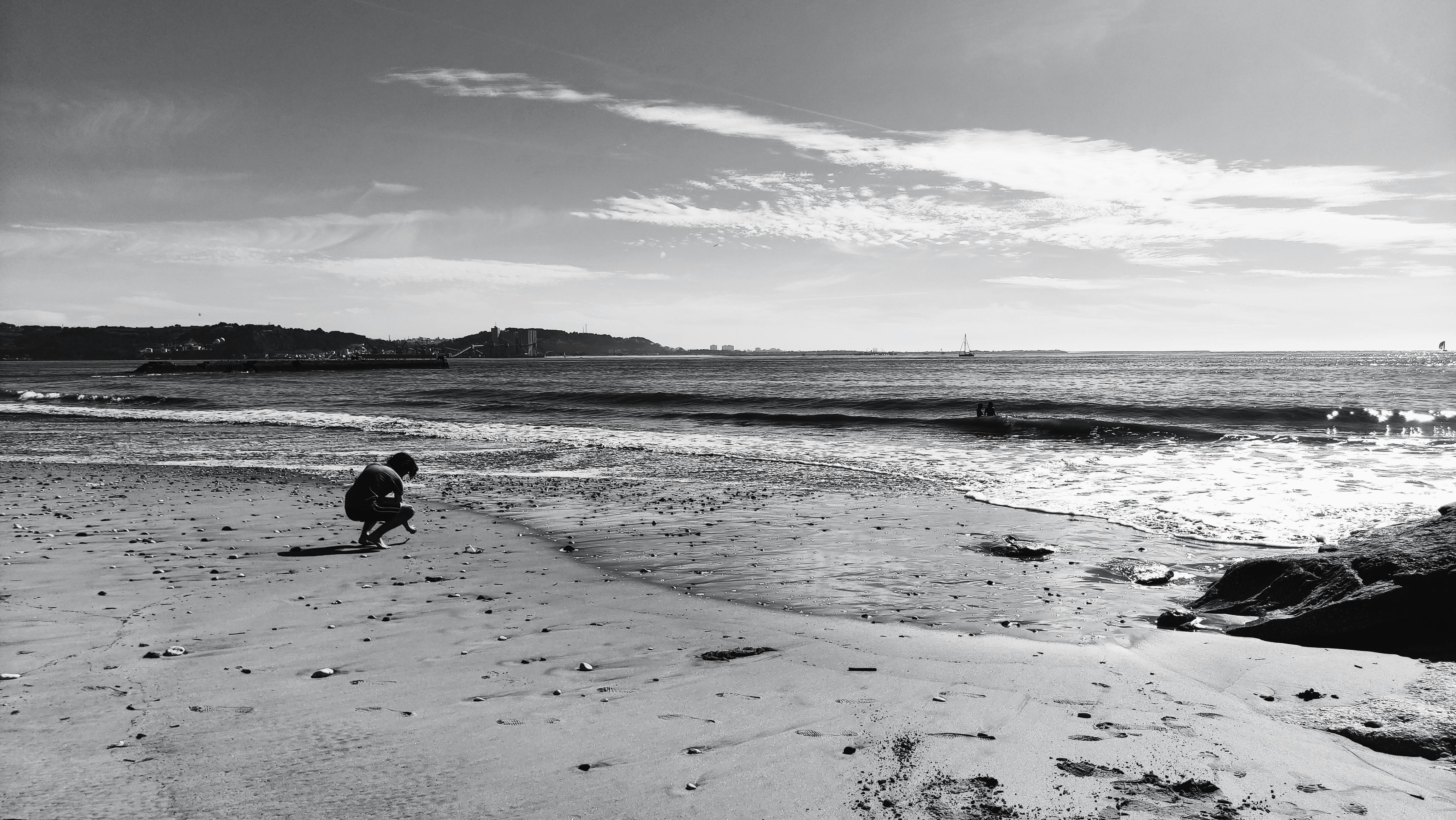 A serene black and white beach scene in Lisbon, Portugal with a person squatting near the shore.
