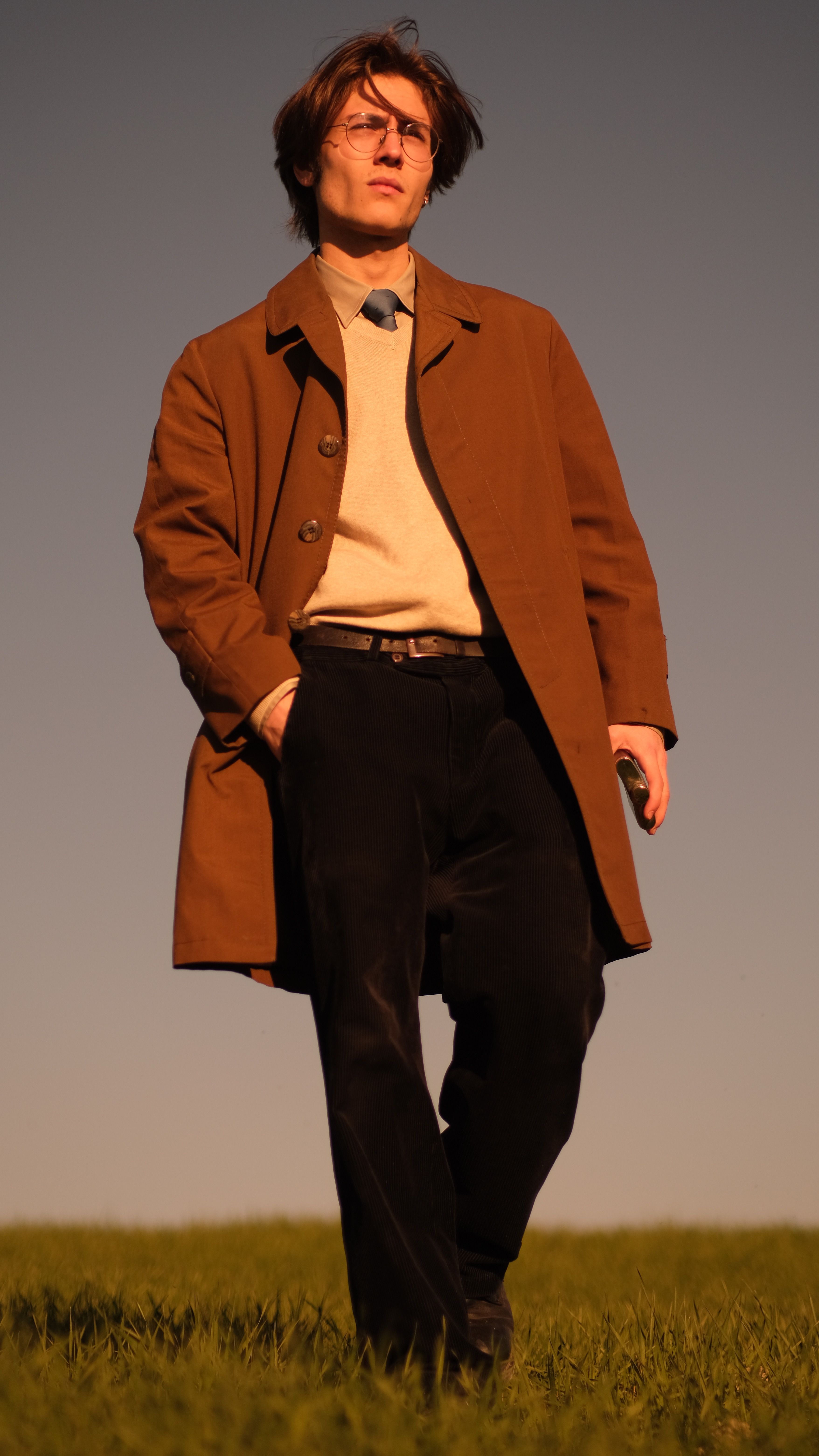Stylish young man in a brown coat standing confidently in a rural landscape.
