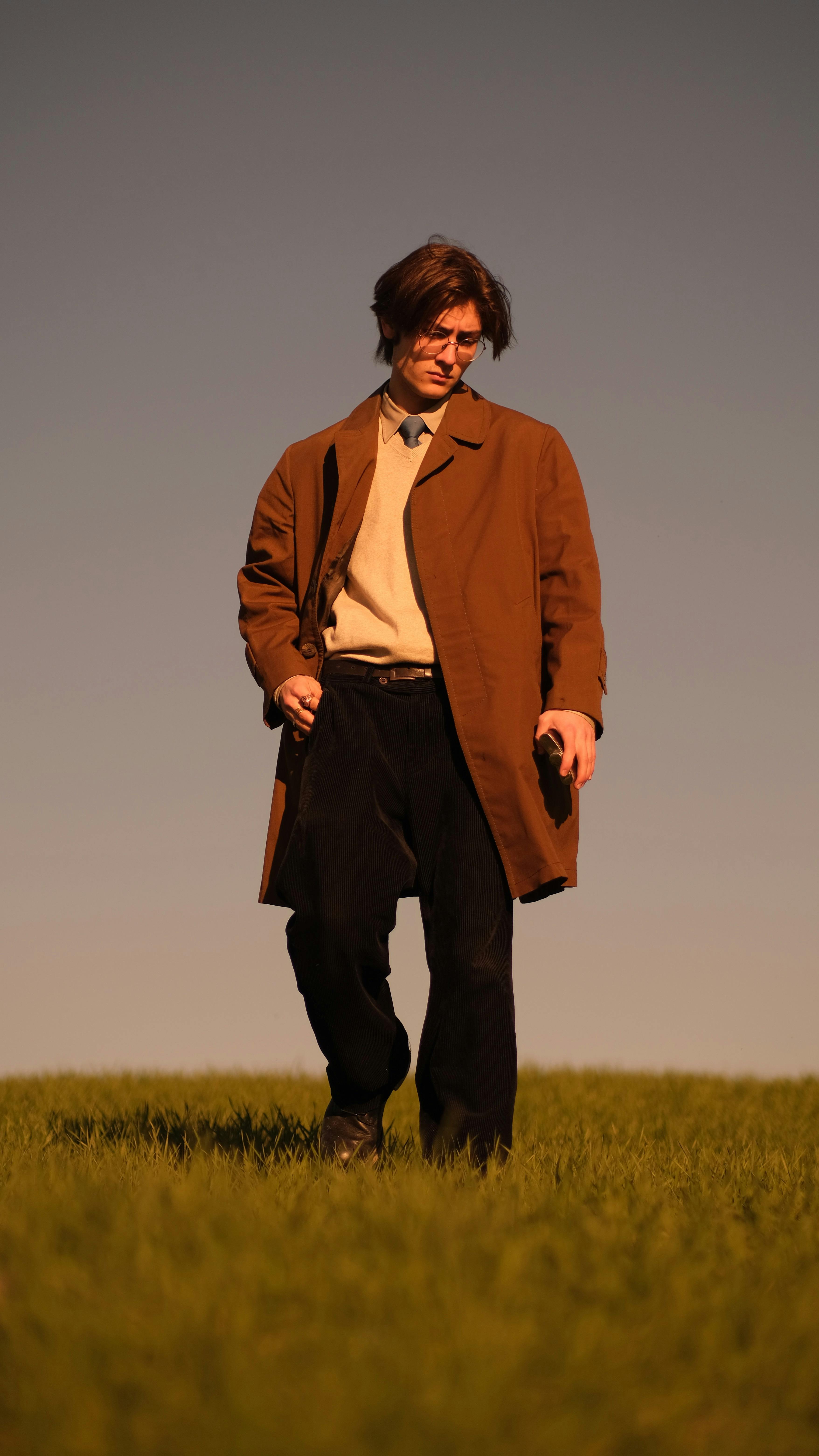 Young man in a trench coat walking on a grassy field under warm sunlight, showcasing fashion style.