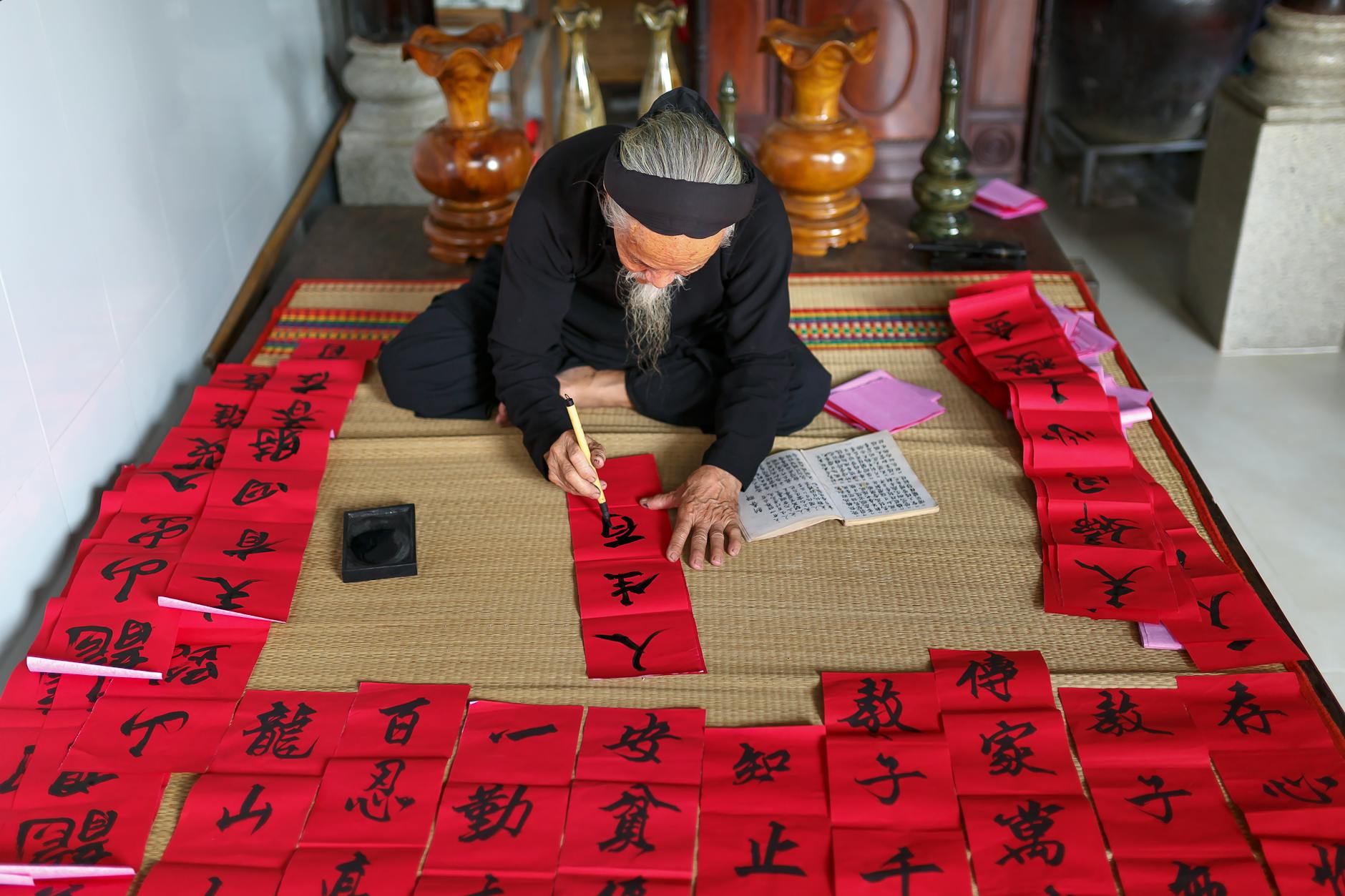 An elderly man practices traditional calligraphy on red paper indoors. Skillful and precise.
