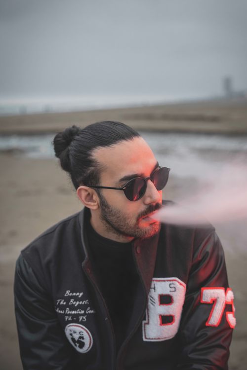 Free Portrait of a stylish man with sunglasses and black hair smoking at a beach in cool weather. Stock Photo