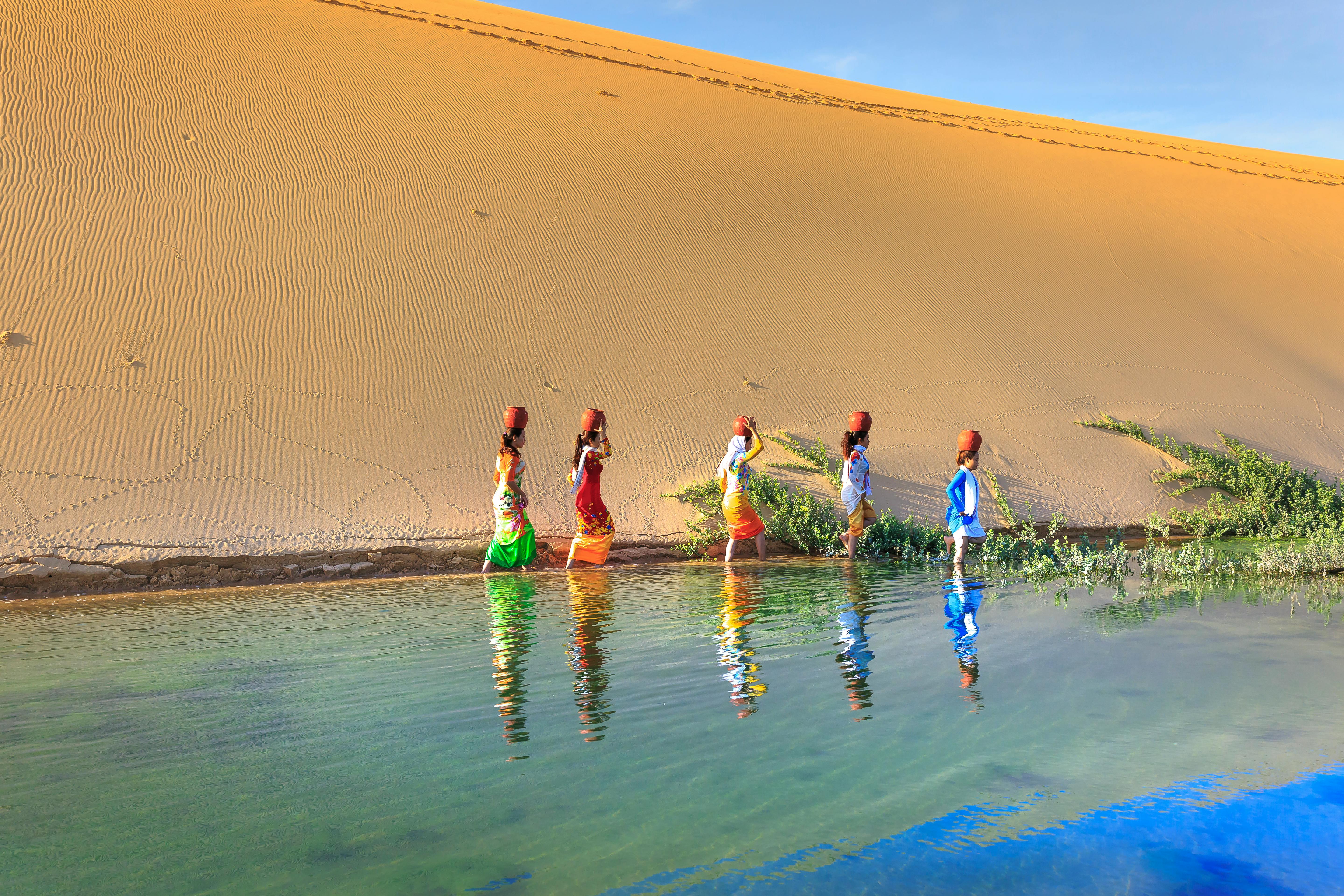 一位色彩缤纷的女性手持水瓶走过沙漠绿洲，倒映出充满活力的身影