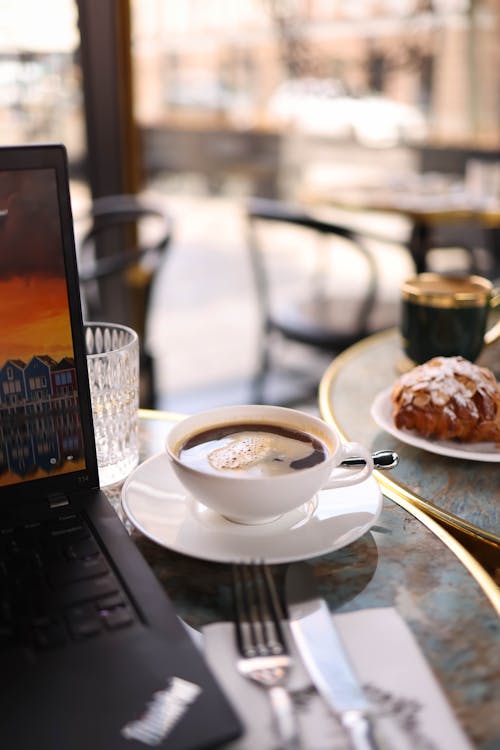 A laptop on a table with a cup of coffee and pastry