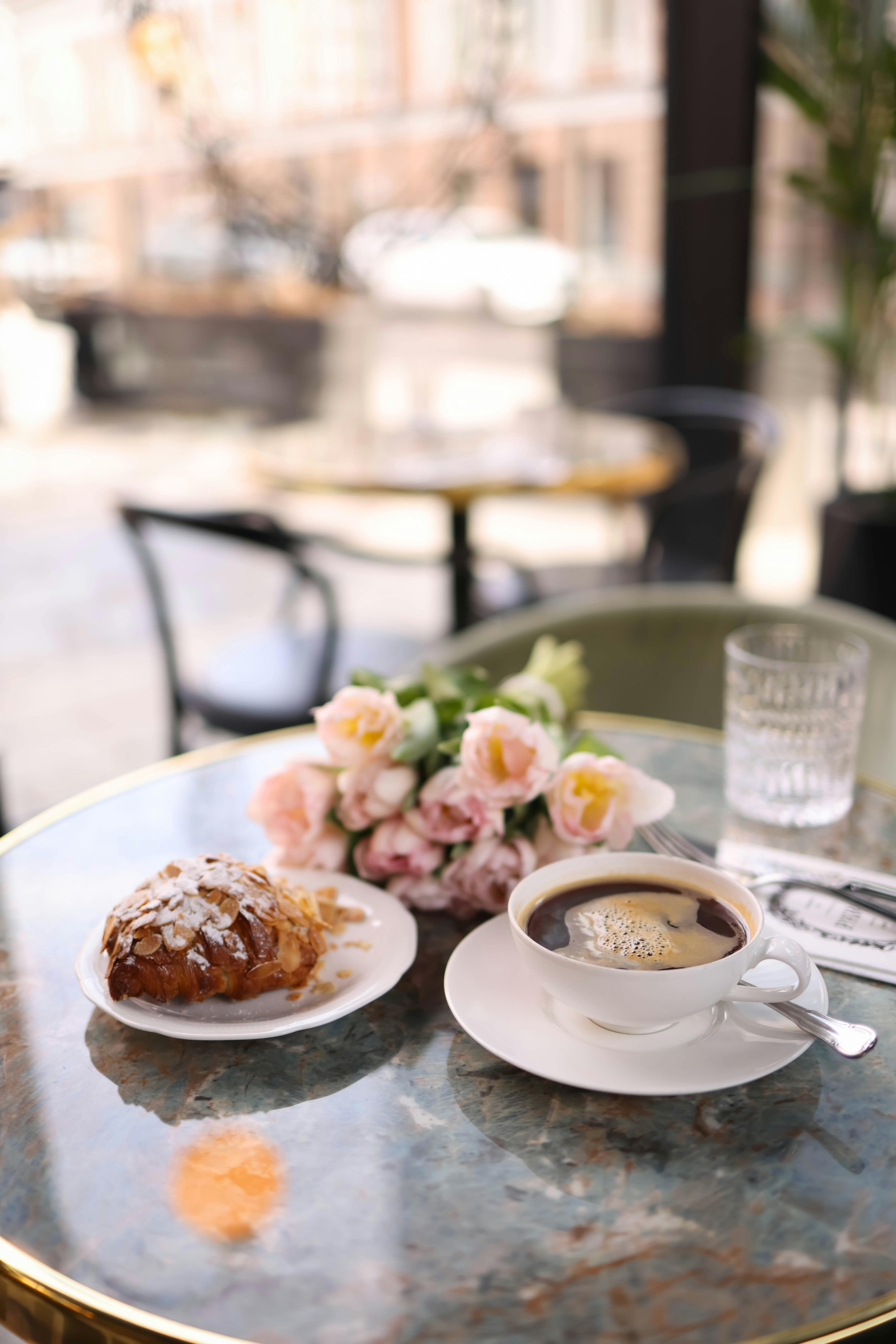 A charming café table with a cup of coffee, pastry, and roses on a sunny day.