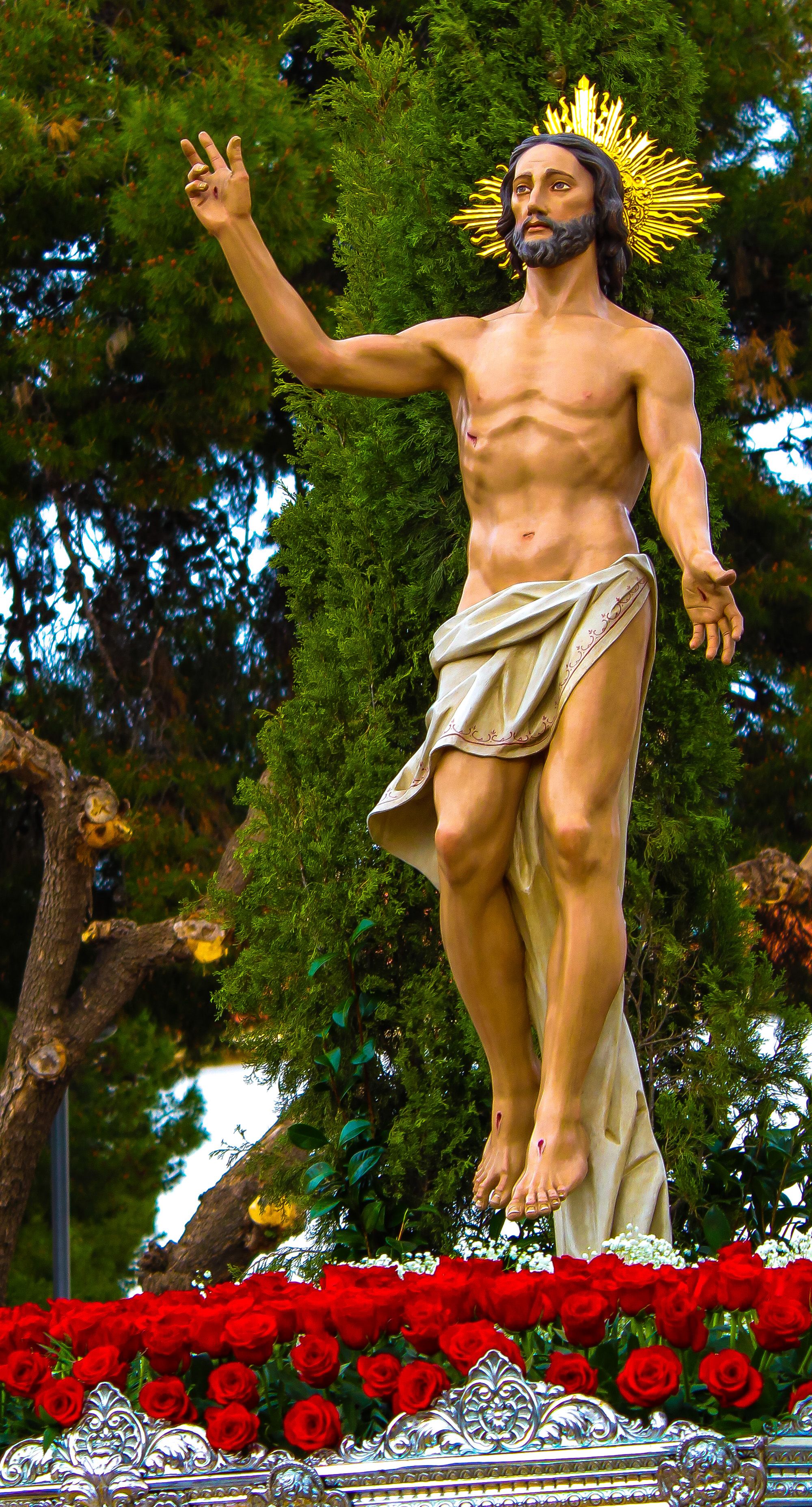 Outdoor statue of Jesus Christ with golden halo surrounded by red flowers, symbolizing spirituality.