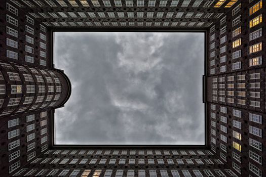 Gazing up through a square opening in a city building complex, revealing a cloudy sky.