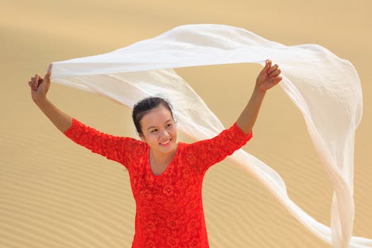 一位身穿红色连衣裙的女子，微笑着在沙丘上摆弄白色布料，尽显幸福与自由