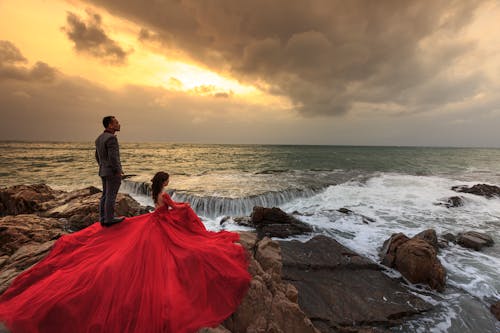 海の前の岩の上に立って座っている赤いガウンの女性と灰色のスーツの男性